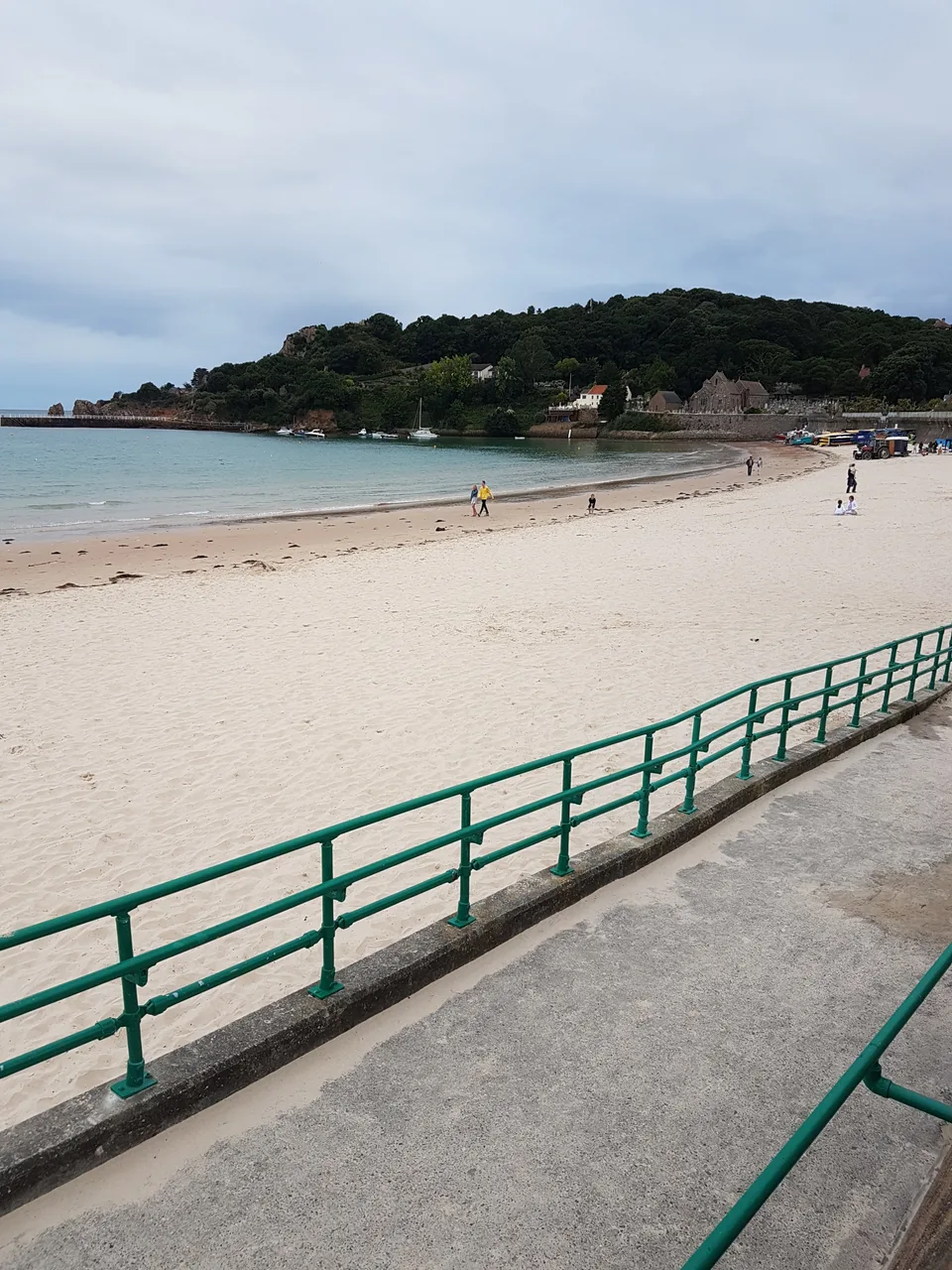 Green railings line a promenade beside pale sand, calm water and a wooded headland at St Brelade's Bay.