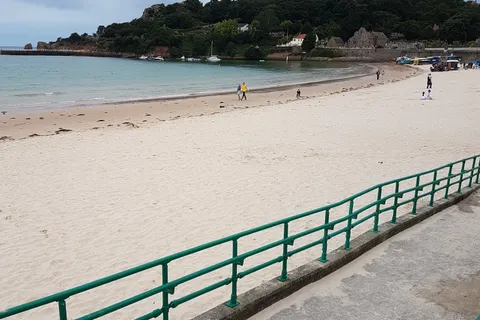 Green railings line a promenade beside pale sand, calm water and a wooded headland at St Brelade's Bay.