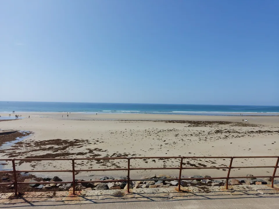Metal promenade railings frame a broad sandy beach with scattered seaweed and the sea beyond at St Ouen's Bay.
