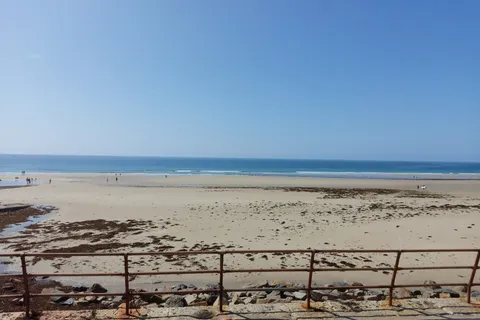 Metal promenade railings frame a broad sandy beach with scattered seaweed and the sea beyond at St Ouen's Bay.