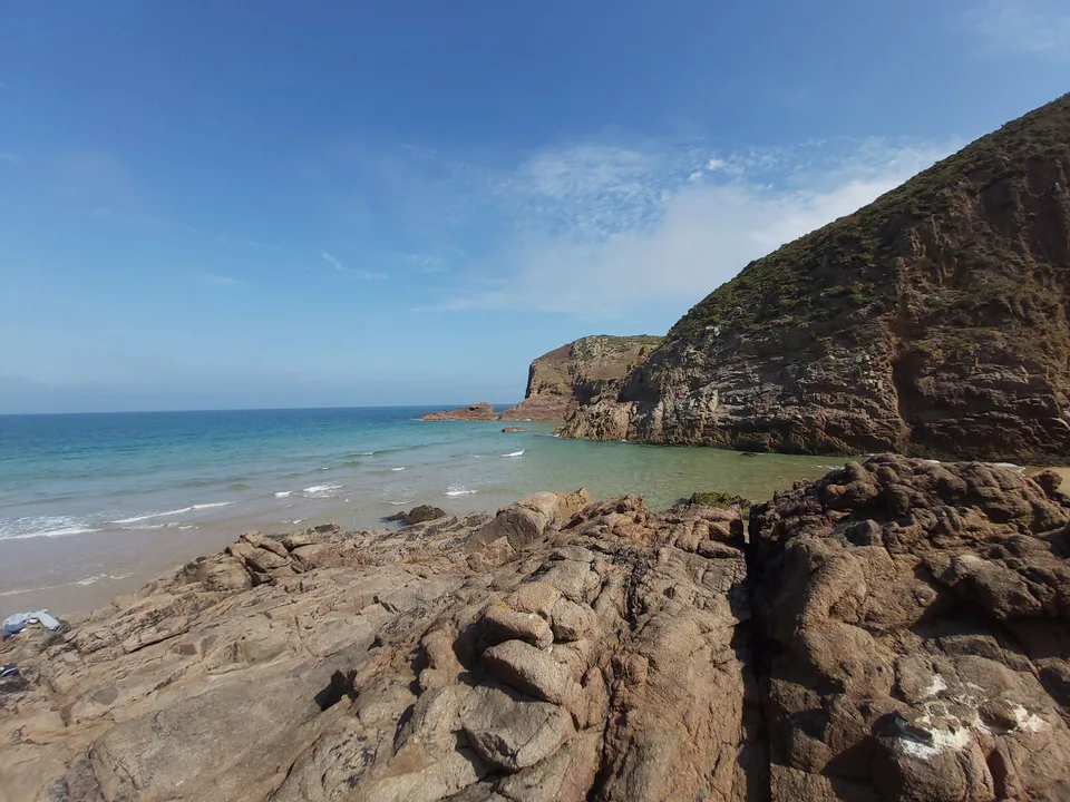 A steep grass-topped cliff borders blue water, with dark shoreline rocks in the foreground at Plémont.