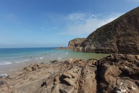 A steep grass-topped cliff borders blue water, with dark shoreline rocks in the foreground at Plémont.