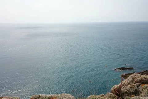 Calm blue water stretches to a hazy horizon beyond foreground grass and a rugged rocky cliff edge.