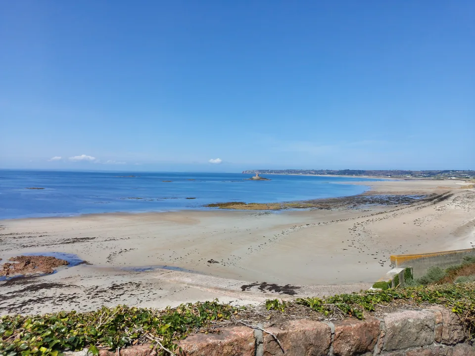 A broad sandy beach and scattered rocks meet clear blue water at St Ouen's Bay under a cloudless sky.