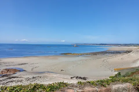 A broad sandy beach and scattered rocks meet clear blue water at St Ouen's Bay under a cloudless sky.