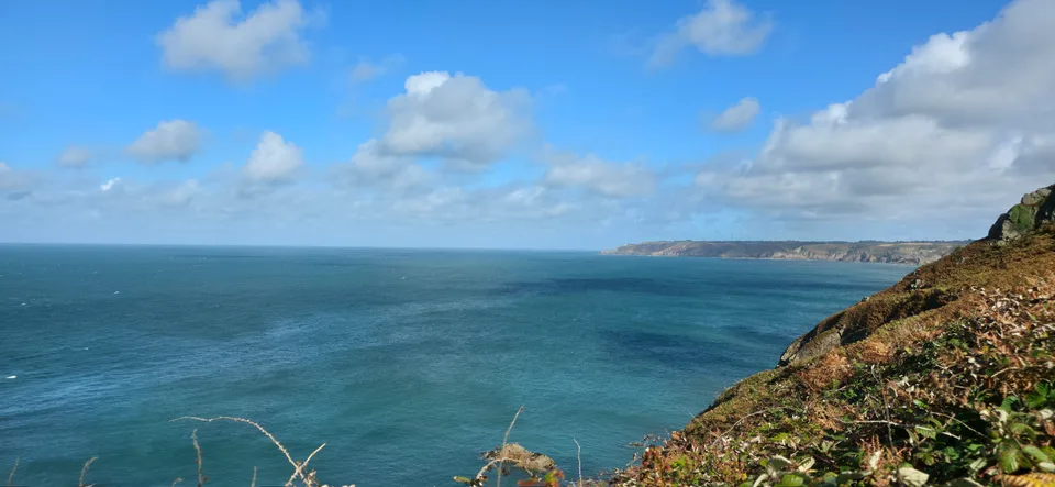 A grassy rocky cliff overlooks blue water and a distant headland near Plémont beneath bright clouds.