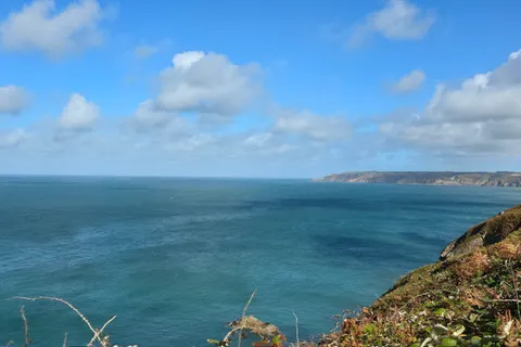 A grassy rocky cliff overlooks blue water and a distant headland near Plémont beneath bright clouds.