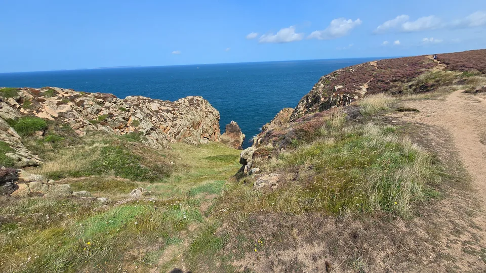 A grassy rocky headland slopes towards blue water at Grosnez with a clear horizon and bright sky.
