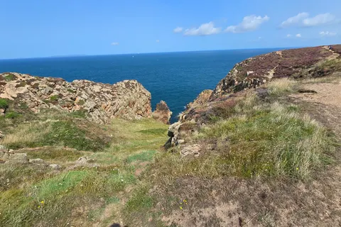A grassy rocky headland slopes towards blue water at Grosnez with a clear horizon and bright sky.