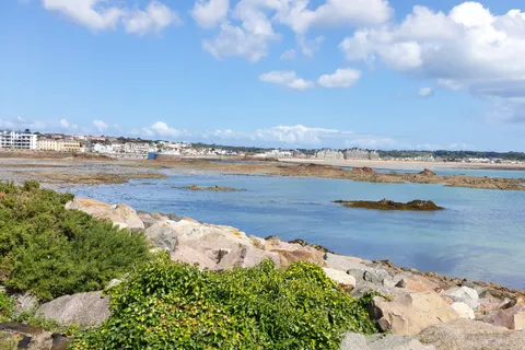 Low green plants grow between pale shoreline rocks beside blue water and white clouds across St Aubin's Bay.