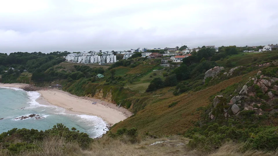 A rocky grassy hillside slopes towards a sheltered sandy bay beneath trees and distant pale buildings.