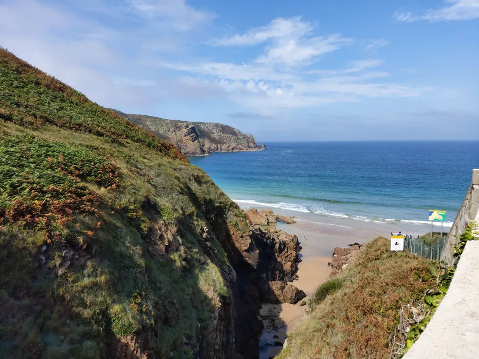 A narrow coastal path beside a steep green slope overlooks Plémont's sandy beach and blue sea.