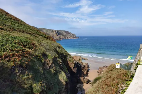A narrow coastal path beside a steep green slope overlooks Plémont's sandy beach and blue sea.