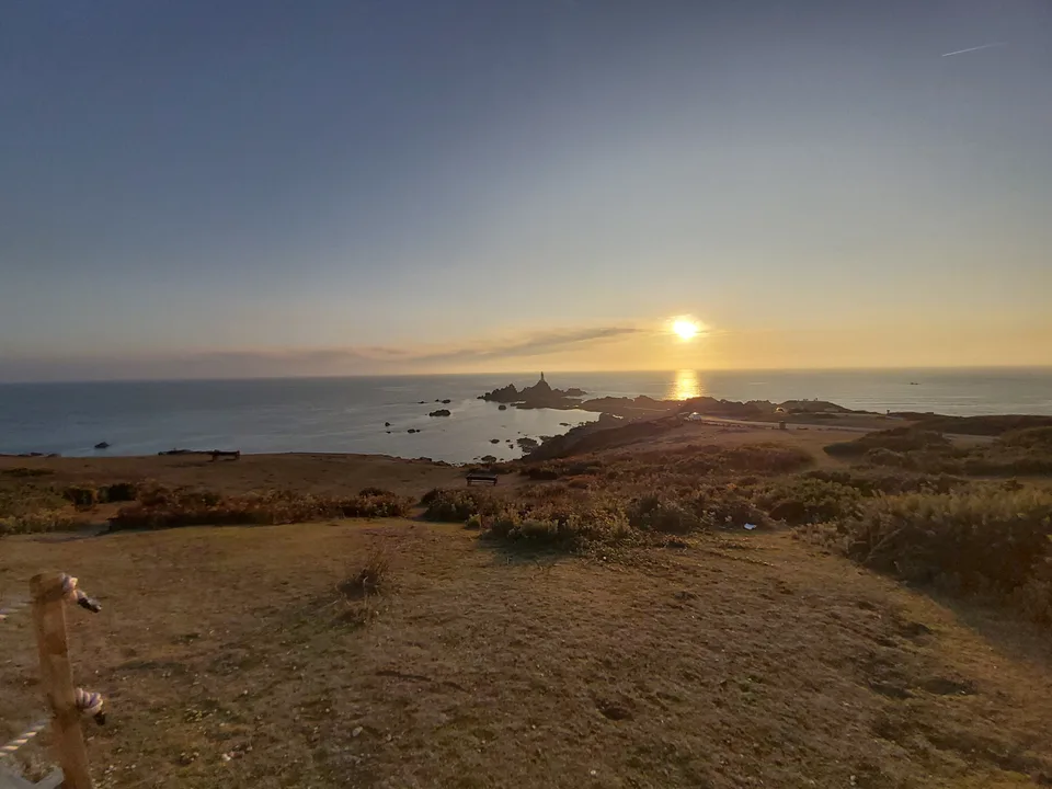 A rough path through coastal grass leads towards dark rocks, La Corbière Lighthouse and a low golden sun.