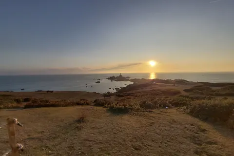 A rough path through coastal grass leads towards dark rocks, La Corbière Lighthouse and a low golden sun.