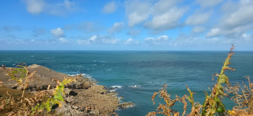 Low coastal plants frame a blue sea and rocky headland at Plémont under a bright clouded sky.