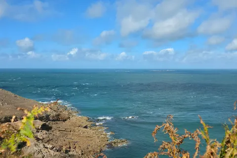 Low coastal plants frame a blue sea and rocky headland at Plémont under a bright clouded sky.