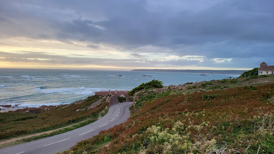 A winding road crosses a grassy coastal headland towards La Corbière Lighthouse beneath a glowing evening sky.