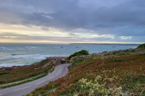 A winding road crosses a grassy coastal headland towards La Corbière Lighthouse beneath a glowing evening sky.