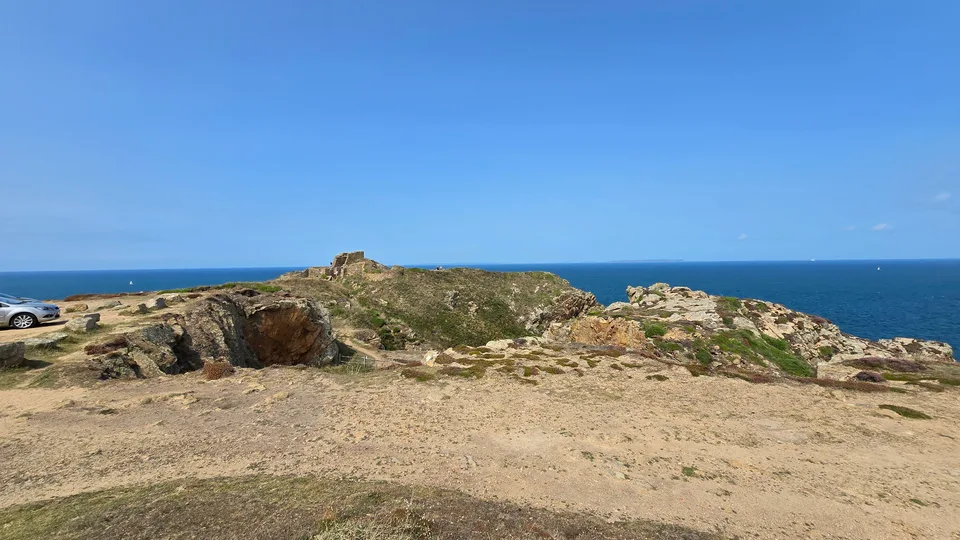 A pale rough path crosses low rocks above the blue sea at Grosnez beneath a clear sky.