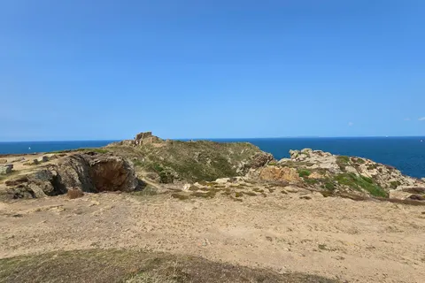 A pale rough path crosses low rocks above the blue sea at Grosnez beneath a clear sky.