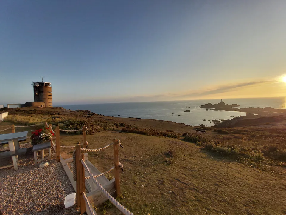 Picnic tables and a rope fence overlook a coastal tower, rocky shore and evening sea at La Corbière.