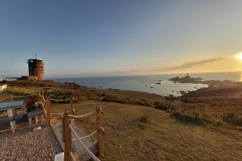 Picnic tables and a rope fence overlook a coastal tower, rocky shore and evening sea at La Corbière.