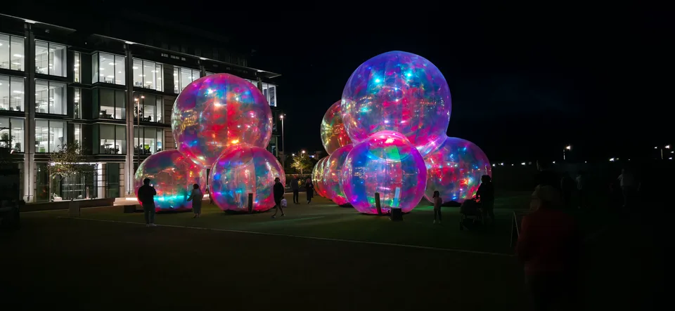 Clusters of large transparent spheres glow with multicoloured reflections outside a lit building at night.