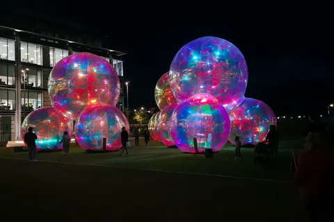 Clusters of large transparent spheres glow with multicoloured reflections outside a lit building at night.
