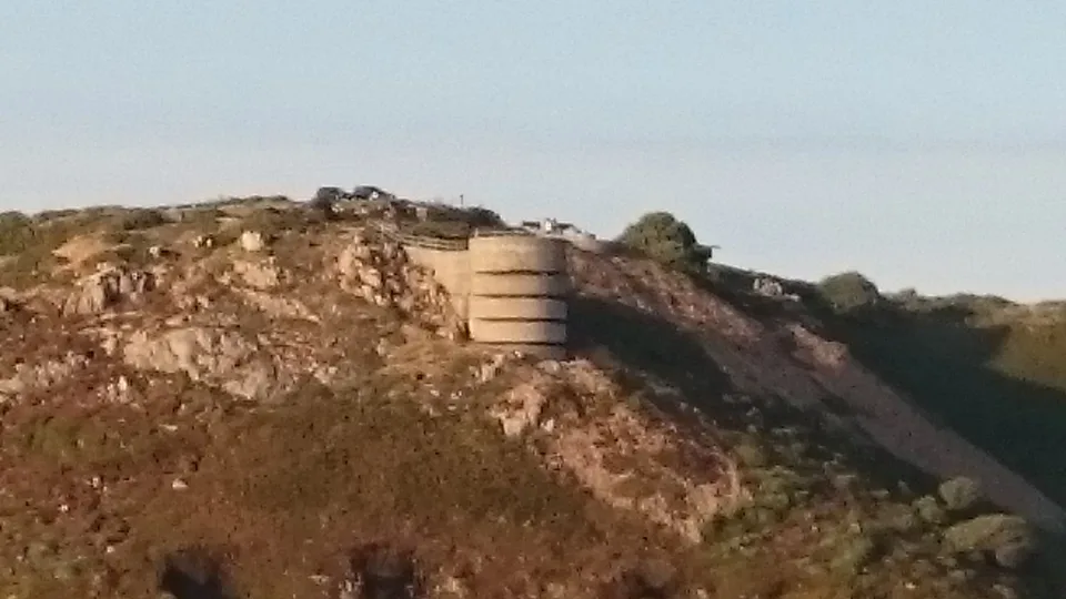 A low concrete structure stands among weathered coastal rocks and grass with the open sea behind.