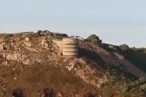 A low concrete structure stands among weathered coastal rocks and grass with the open sea behind.