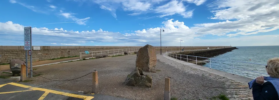 A road sign and stone paving mark the entrance to St Catherine's Breakwater beside blue sea and scattered clouds.