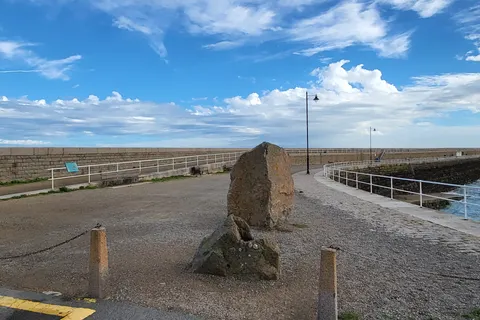 A road sign and stone paving mark the entrance to St Catherine's Breakwater beside blue sea and scattered clouds.