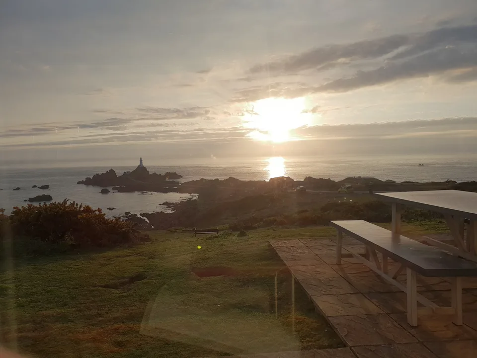 Low sunlight reflects on the sea beside La Corbière Lighthouse, with a grassy slope and picnic table in front.