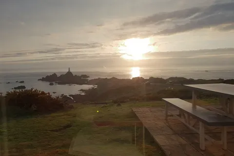 Low sunlight reflects on the sea beside La Corbière Lighthouse, with a grassy slope and picnic table in front.