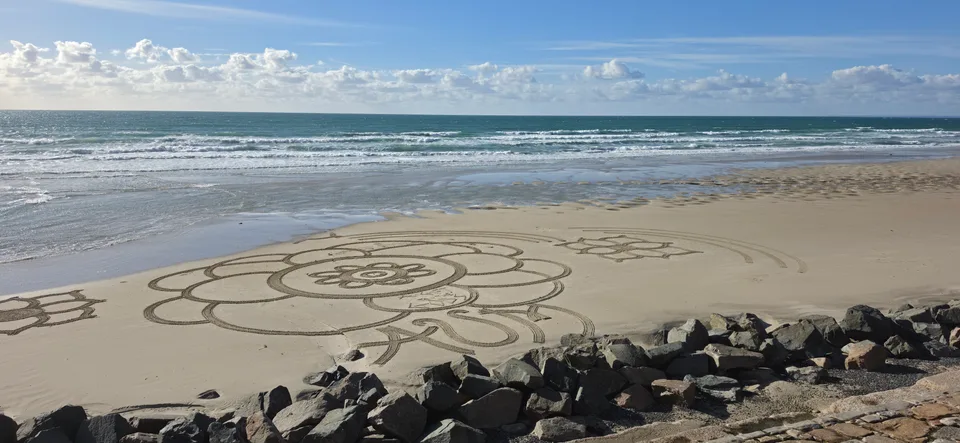 A large flower-shaped drawing fills the sand beside a stone seawall and breaking waves at St Ouen's Bay.