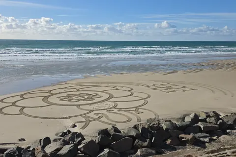A large flower-shaped drawing fills the sand beside a stone seawall and breaking waves at St Ouen's Bay.