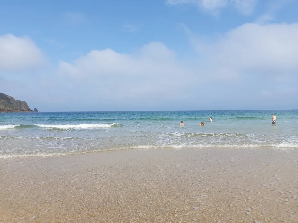Small waves wash over smooth wet sand at Plémont beneath blue sky and scattered clouds.