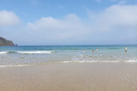 Small waves wash over smooth wet sand at Plémont beneath blue sky and scattered clouds.