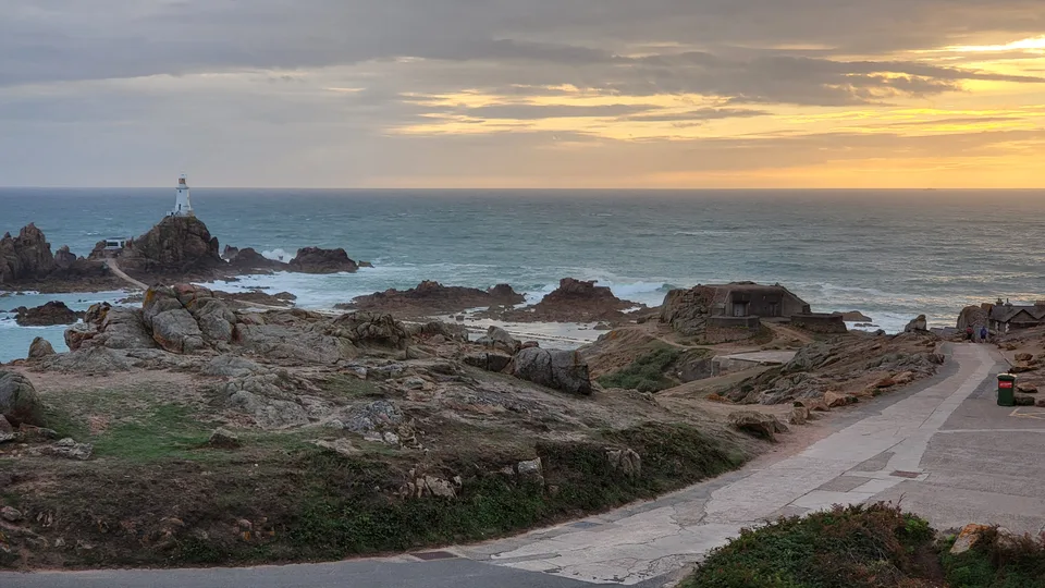 A causeway crosses rugged rocks towards La Corbière Lighthouse below a band of golden evening cloud.