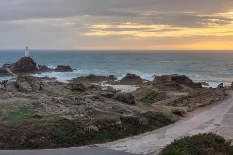 A causeway crosses rugged rocks towards La Corbière Lighthouse below a band of golden evening cloud.