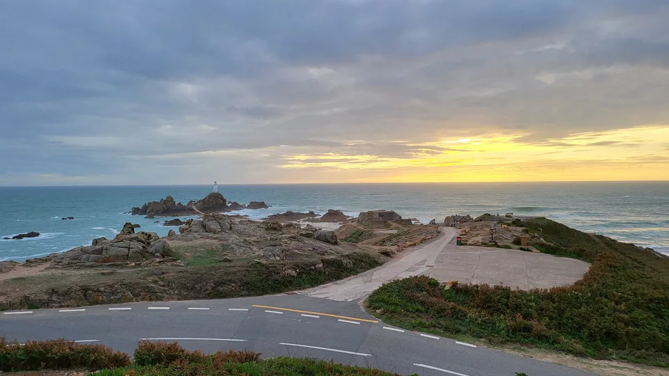 A curving coastal road and rocky headland frame La Corbière Lighthouse beneath a golden horizon and grey clouds.