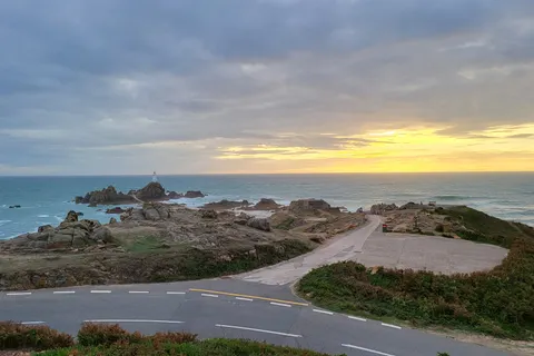 A curving coastal road and rocky headland frame La Corbière Lighthouse beneath a golden horizon and grey clouds.
