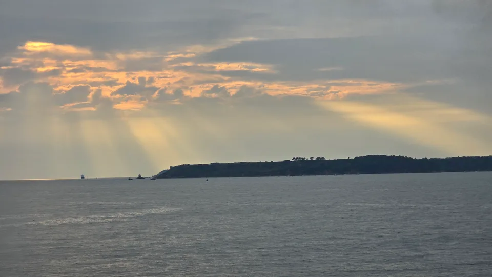 Golden sunbeams spread through grey clouds above open water and a distant headland in St Aubin's Bay.