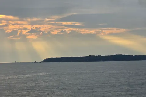 Golden sunbeams spread through grey clouds above open water and a distant headland in St Aubin's Bay.