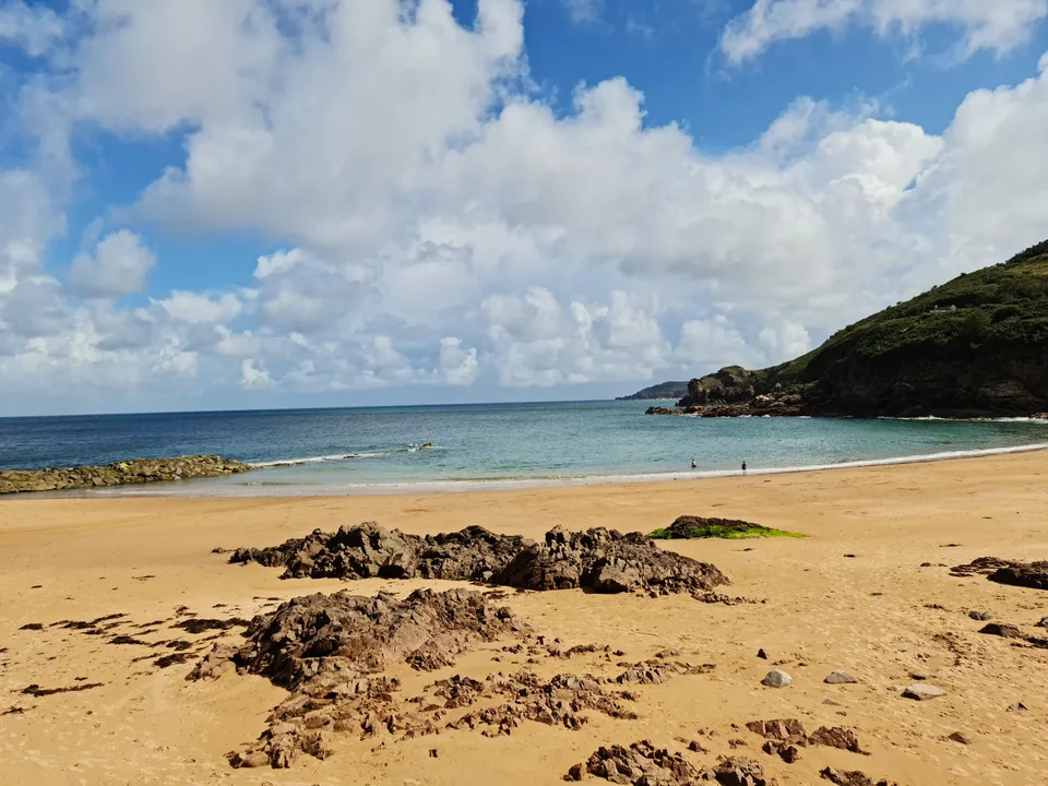 Golden sand and exposed rocks frame clear blue water and a grassy headland at Grève de Lecq beneath large clouds.