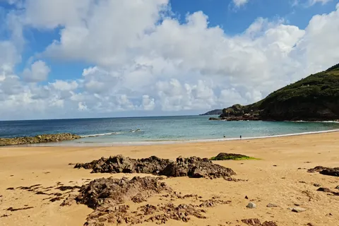 Golden sand and exposed rocks frame clear blue water and a grassy headland at Grève de Lecq beneath large clouds.