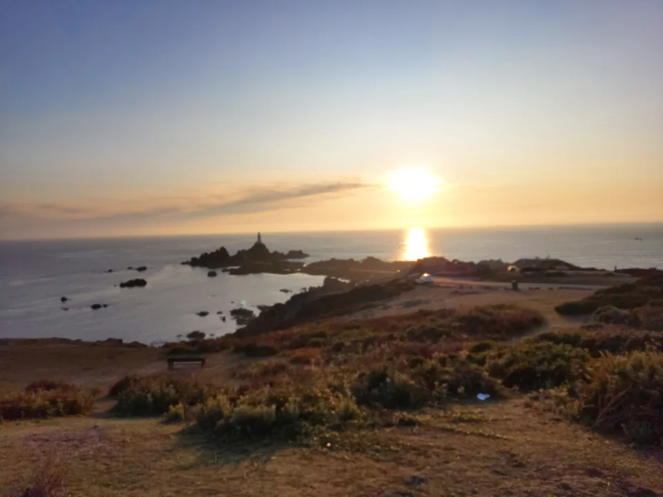 A golden sun reflects across the sea beside La Corbière Lighthouse above a dark grassy coastal foreground.