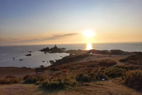 A golden sun reflects across the sea beside La Corbière Lighthouse above a dark grassy coastal foreground.
