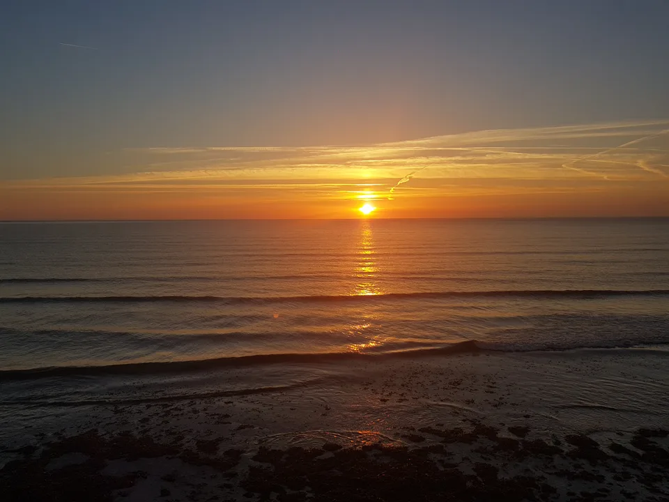 A low golden sun casts a bright reflection across the sea above small waves and a dark shoreline.